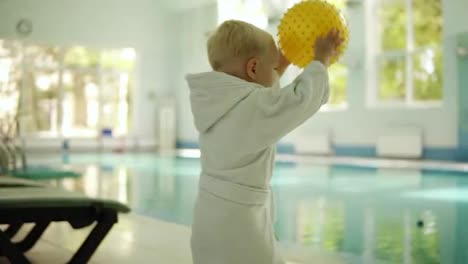 Cute-little-boy-throwing-a-yellow-ball-to-the-pool-at-the-leisure-center.-Swimming-pool-indoors.-Boy-wears-small-white-terry-bathrobe.-Slow-motion