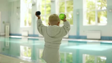 Backside-view-of-a-little-boy-in-bathrobe-throwing-balls-from-both-hands-into-the-swimming-pool.-Playful.-Outside-the-pool.-Pure-water