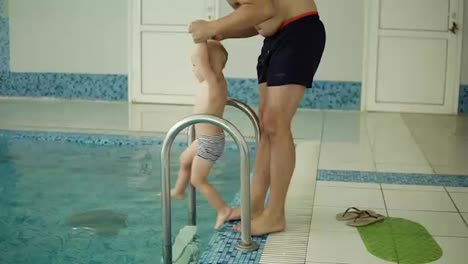 Parents-teaching-their-son-to-swim-in-the-pool.-Young-family-in-the-pool-teaching-his-son-to-swim.-Father-outside-the-pool,-puts-child-into-water,-mother-helping-him-to-swim.-Indoors.-Slow-motion