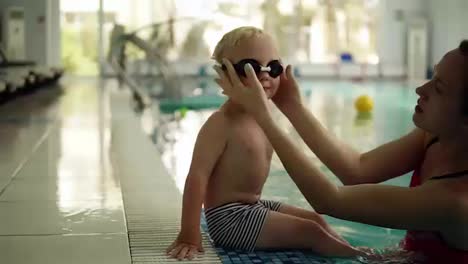 A-small-swimmer-prepares-to-swim.-The-boy-sits-on-the-edge-of-the-pool,-his-mother-puts-on-his-glasses-for-swimming.-Waving-his-feet-on-the-water.-Indoors