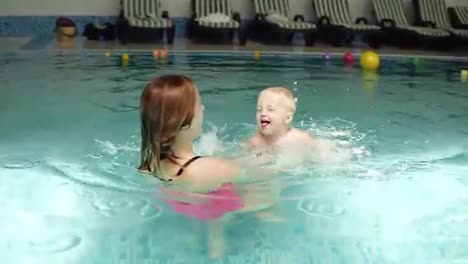 Cute-baby-having-swimming-lesson-with-his-mother.-Healthy-family-teaching-their-baby-to-swim-in-the-swimming-pool.-Young-mother-takes-his-son-and-embraces-him-while-the-kid-is-smiling