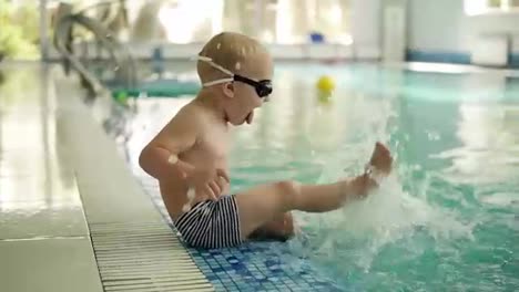 A-small-blond-boy-sits-on-the-edge-of-the-pool-in-swimming-trunks-and-glasses-and-sprinkles-water-with-his-feet.-Splashing-the-water.-Having-fun.-Indoors