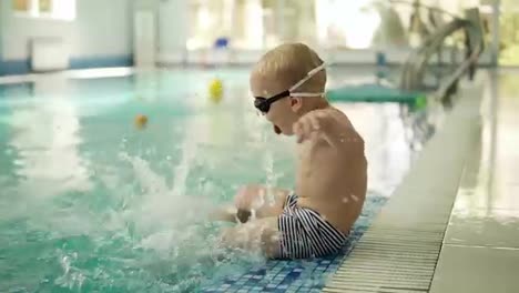 A-little-boy-with-blond-hair-sits-by-the-pool-with-his-feet-in-the-water.-Dabbles,-splashes-water-with-his-feet.-Side-view.-Hotel-swimming-pool.-Slow-motion
