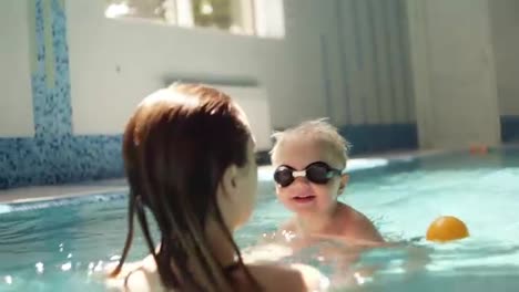 Young-woman-holding-his-son,-teaching-to-swim.-Supporting-above-the-water.-Indoors-swimming-pool.-Little-boy-in-swimming-glasses