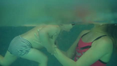 Side-view,-an-underwater-shot-of-young-woman-in-pink-swimming-suit-supporting-his-son-in-water-pool.-Teaching-how-to-swim.-Indoors-swimming-pool