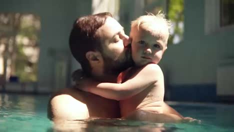 Portrait-of-a-father-and-son-spending-time-together-in-the-swimming-pool.-Sunrays-laying-on-them.-Father-is-hugging-his-son,-embracing,-kissing.-Little-boy-is-on-fathers-hands.-Indoors-swimming-pool