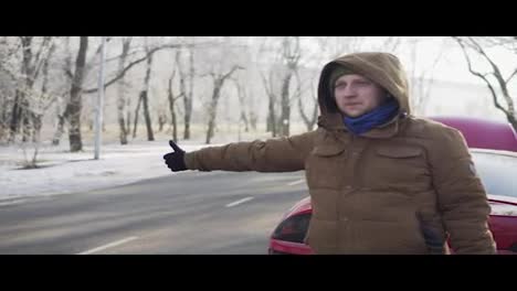 Attractive-young-man-hitchhiking-on-a-road-with-his-broken-down-car-behind-him