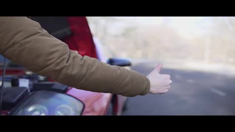 Closeup-hand-of-a-man-hitchhiking-on-a-road-with-his-broken-down-car-close-to-him