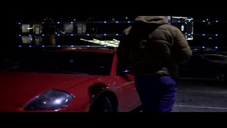 Man-getting-into-red-sport-car-on-drivers-side.-City-lights-at-night-on-the-background