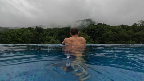 A-Muscular-Man-Walks-Inside-Infinity-Pool-With-Lush-Mountain-View-on-Rainy-Day