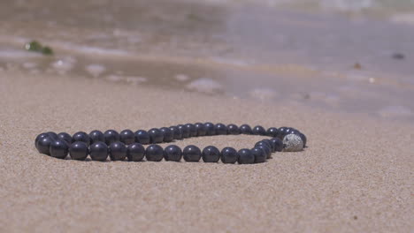 Cinematic-shot-of-a-pearl-nacklace-with-silver-jewellery-laying-on-a-sand-beach-close-to-the-ocean,-120-FPS,-Slomo