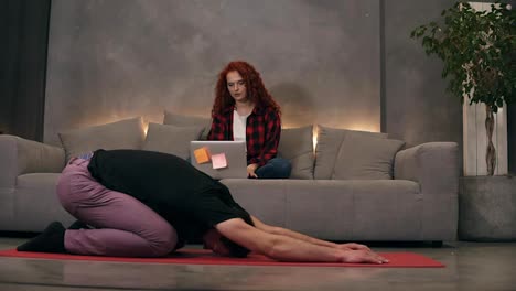 Young-caucasian-couple-at-relaxing-home-living-room,-curly-readheaded-firl-using-modern-wireless-laptop-on-a-grey-couch-while-her-boyfriend-is-doing-yoga,-stretching-on-a-red-mat.-Front-view.-Loft-designed-living-room