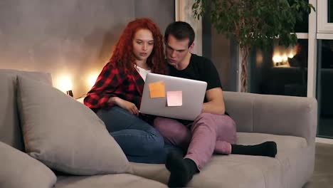Beautiful-young-couple-sitting-together-with-grey-laptop-on-couch.-Couple---shorthaired-man-and-red-headed-girl-watching-something-on-laptop-sitting-on-sofa-in-stylish-loft-living-room-in-the-evening