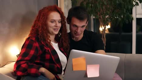 Beautiful-young-couple-sitting-together-with-grey-laptop-on-couch.-Couple---shorthaired-man-and-red-headed-girl-watching-together-something-funny-on-laptop.-Talking-and-smiling.-Sitting-on-sofa-in-stylish-loft-living-room-in-the-evening.-Close-up