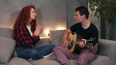A-husband-is-singing-a-song-and-playing-a-guitar-for-his-wife-who-is-also-sitting-with-him-on-a-sofa-and-sing-with-him.-She-likes-to-watch-him-perform.-Taking-time-together-at-home-at-living-room