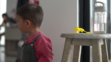 Kleiner-Hispanischer-Lateinamerikanischer-Junge,-Der-Mit-Einem-Gelben-Kaputten-Plastikfroschspielzeug-Auf-Einem-Holzhocker-Spielt-Und-Wegen-Staub-In-Der-Luft-Niest