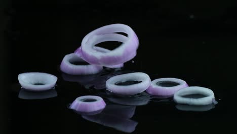 Zeitlupenaufnahme-Von-Weißen-Und-Violetten-Zwiebelringen,-Die-Auf-Eine-Schwarze,-Mit-Wasser-Bedeckte-Oberfläche-Fallen