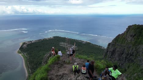 People-standing-on-a-mountain-peak-above-ocean-peninsula,-aerial-view