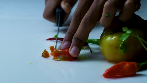 Slow-Motion-Shot-of-a-Man-Cutting-a-Red-Bombay-Chili-Pepper-on-top-of-a-White-Surface,-next-to-other-Vegetables