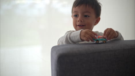 Close-up-of-a-young-hispanic-boy-playing-with-a-blue-car-toy-on-a-grey-couch-wearing-pajamas-on-a-cozy-morning