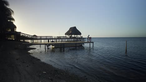 Dusk-at-Punta-Gorda-beach-as-the-silhouette-of-a-pair-of-kids-play-at-the-edge-of-the-pier-in-Roatan,-Honduras---Handheld