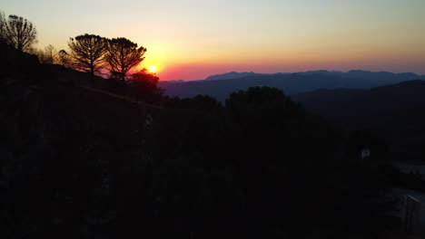 Eine-Drohne-Schiebt-Sich-Bei-Sonnenuntergang-Durch-Die-Silhouetten-Der-Bäume-Und-Berge