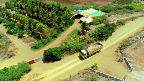 4k-aerial-cinematic-tilt-down-of-yellow-dump-truck-filled-with-dirt-and-rubbish-driving-through-rural-farmland-with-windpumps-on-a-dirt-road-during-a-beautiful-sunny-day-in-the-Caribbean,-Curacao