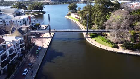 Drone-Aerial-View-zooming-backwards-over-people-walking-along-Suspension-pedestrian-bridge-in-Claisebrook-Cove,-East-Perth-and-Swan-River