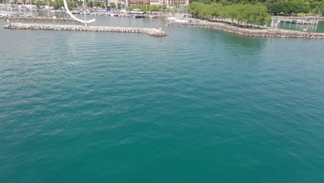 Drone-flying-over-beautiful-teal-lake,-tilting-up-and-revealing-a-yacht-harbor-in-Lausanne,-Switzerland