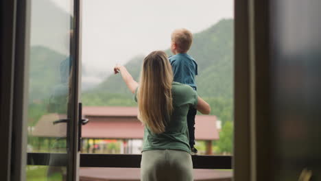 Careful-mother-shows-green-hills-holding-son-on-balcony