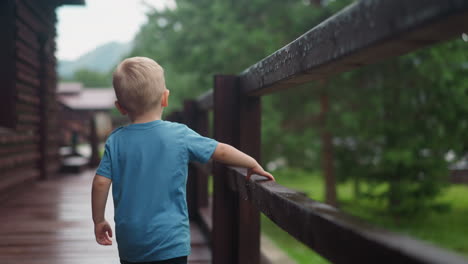 Little-blond-child-walks-touching-wet-wooden-railing-on-deck