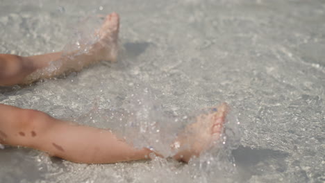 Kleines-Kind-Schwingt-Beine-Und-Spritzt-Klares-Wasser-Am-Strand