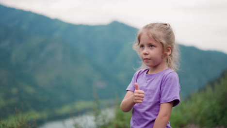 Cute-little-girl-shows-thumb-up-in-valley-against-mountains