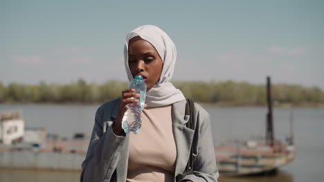 Müde-Muslimische-Frau-öffnet-Flasche-Wasser-Am-Flussufer