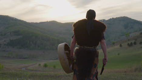 Actor-walks-carrying-shamanic-drum-against-old-hills