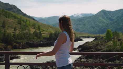 Woman-looks-around-on-bridge-over-river-in-mountain-valley
