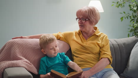 Funny-little-boy-turns-book-pages-with-granny-in-living-room