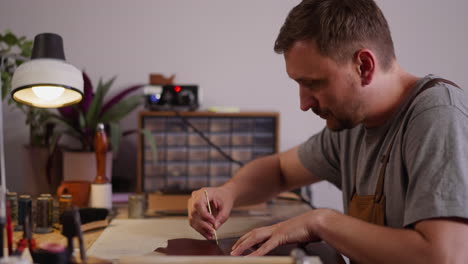 Concentrated-man-cuts-leather-with-small-knife-at-workplace