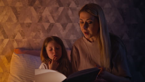 Little-girl-listens-to-mom-reading-book-in-dark-bedroom
