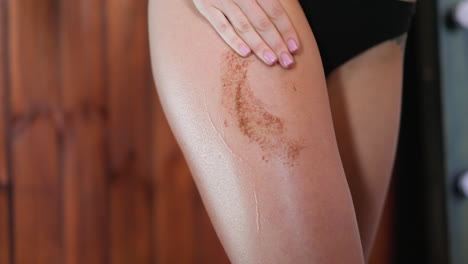 Young-woman-spreads-coffee-scrub-on-thigh-making-massage
