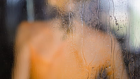 Woman-takes-shower-behind-water-drops-flowing-down-on-glass