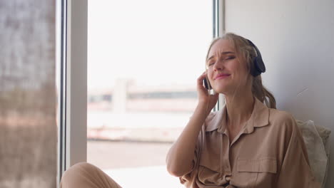 Young-woman-enjoys-music-in-headphones-sitting-on-windowsill
