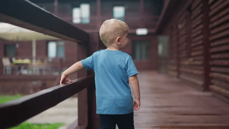 Sweet-boy-walks-holding-handrail-along-wet-veranda-deck