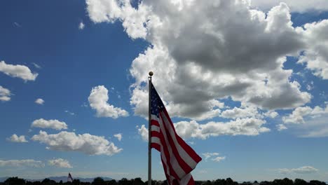 American-flag-USA-blowing-waving-in-the-wind-on-beautiful-sunny-summer-day-with-clouds-and-blue-skies-as-drone-flys-around-flagpole-with-small-town-cresting-in-bottom-of-shot---in-4K-60fps
