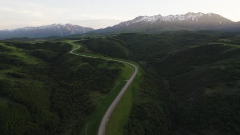 Winding-road,-snow-capped-mountain-peaks-at-sunset,-aerial-panoramic-view