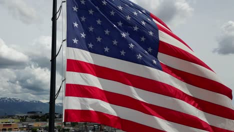 American-flag-USA-blowing-waving-in-the-wind-on-beautiful-sunny-summer-day-with-lots-of-puffy-white-clouds-and-blue-skies-overlooking-Wasatch-Mountains-as-drone-flies-around-flag-pole---in-4K-60fps