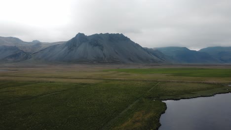 Drone-Estático-Disparó-Sobre-Un-Campo-Verde-Debajo-De-Enormes-Picos-Islandeses-Y-Niebla