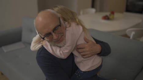 Little-granddaughter-hug-old-elder-grandfather-at-home