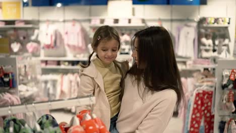 Schöne-Mutter-Schaut-Kleidung-Auf-Kleiderbügel-Mit-Tochter-Im-Einkaufszentrum