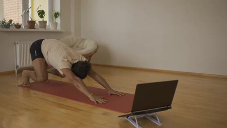 Active-bearded-man-doing-yoga-following-instruction-video-online-trainer-on-laptop-at-home